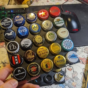 May include: A collection of colorful beer bottle caps, featuring various brand logos and designs, arranged in a black holder. The caps are held by a hand, with a glass of beer and a computer setup in the background.