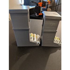 May include: Two gray 3D printed battery organizers with multiple compartments. Each organizer holds several AA batteries, labeled "EBL 2000".