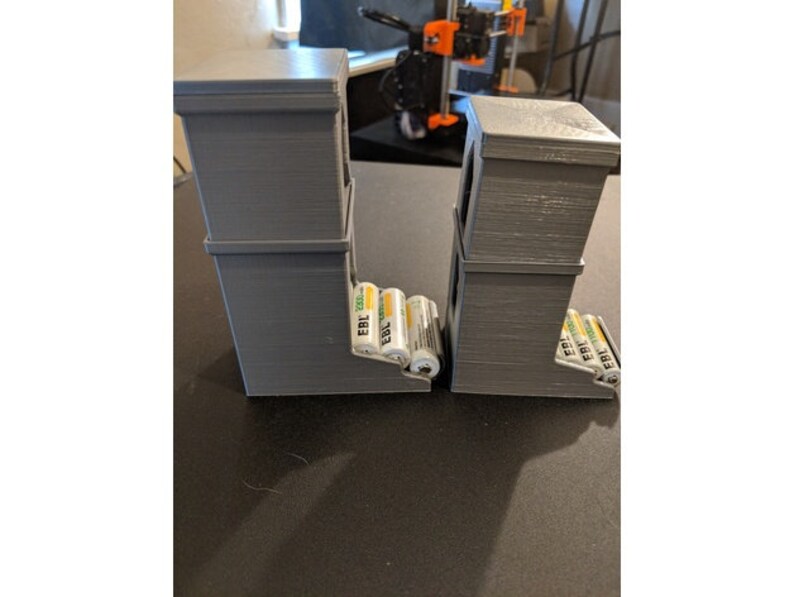May include: Two gray 3D printed battery organizers with multiple compartments. Each organizer holds several AA batteries, labeled "EBL 2000".