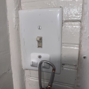 Könnte beinhalten: Ein weißer Lichtschalter mit einem einzigen Kippschalter und einer weißen Steckdose darunter. Ein silberner Karabiner mit Schlüsseln hängt an der Steckdose.