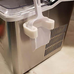 Puede incluir: Máquina de hacer hielo de acero inoxidable con una cuchara y un soporte blancos en el lateral. La máquina de hielo tiene una tapa transparente y una rejilla de ventilación en el lateral. La cuchara encaja en el soporte.