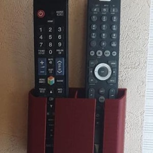 Puede incluir: Un soporte de plástico rojo para mandos a distancia con dos compartimentos, que contiene dos mandos a distancia negros. Los mandos a distancia tienen numerosos botones y están diseñados para un televisor o un dispositivo similar. El soporte está montado en una pared texturizada.