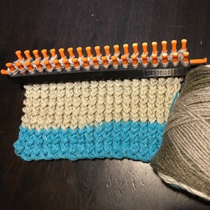 May include: A knitting loom with beige and teal yarn, creating a striped knitted fabric. The loom has orange pegs and a black base.