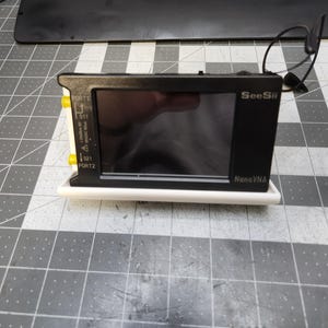 Peut inclure: Un appareil SeeSii NanoVNA noir avec un écran, des ports jaunes et une base blanche. L'appareil est équipé d'une antenne et se trouve sur une surface grise avec un motif quadrillé. L'écran affiche une réflexion sombre.