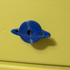 Op de afbeelding: Een blauwe 3D-geprinte Saturnus-vormige ladegreep. De greep heeft een ring om de planeet.