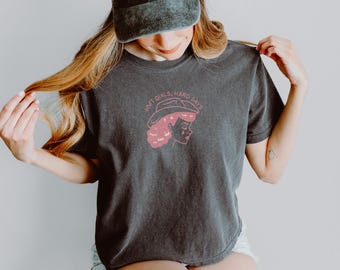 Könnte beinhalten: Dunkelgraues T-Shirt mit einer rosa Grafik einer Frau mit Cowboyhut und dem Text "SOFT GIRLS, HARD DAYS". Eine dunkelgraue Baseballkappe mit dem Wort "HOWDY" in Rosa wird auf dem Kopf getragen.