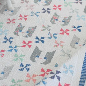 Op de afbeelding: Een witte quilt met een herhalend patroon van katsilhouetten en windmolenblokken in diverse tinten roze, blauw en grijs. De stof bevat bloemen-, stippen- en streepmotieven, afgewerkt met een blauw-witte gestreepte rand.