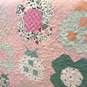 May include: Close-up of a patchwork quilt with a pink background. The quilt features floral and geometric patterns in shades of pink, green, white, and red. The quilt has a quilted texture with a floral design.