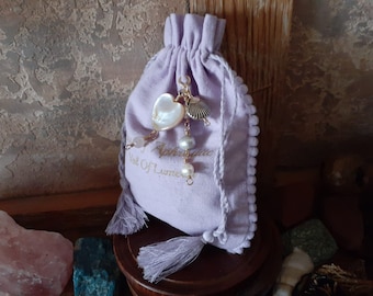 May include: A lavender drawstring pouch with a white heart charm, a seashell charm, and white pearl beads. The pouch has a tassel and the text "West Of Luna" is embroidered on the front.