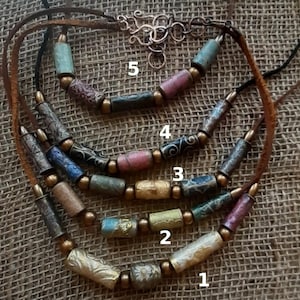 May include: Five strands of a necklace with brown leather cord and gold metal clasps. Each strand features different colored and patterned cylindrical beads. The beads are made of paper and have a rustic, vintage look.
