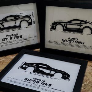 May include: Three framed automotive art pieces. Each features a black outline of a car model against a white background. The frames display the names: Nissan GT-R R35, Ford Mustang, and Toyota Supra MK5.