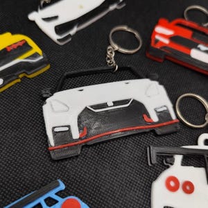 May include: A collection of car-shaped keychains in various colors, including white, red, yellow, and blue. Each keychain features a detailed car design with a metal keyring attached. The keychains are made of plastic.
