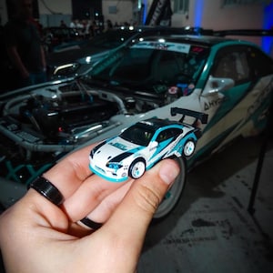 Può includere: Una piccola auto giocattolo bianca e blu con dettagli neri e uno spoiler posteriore. L'auto giocattolo è tenuta davanti a un'auto più grande, abbinata, con un design turchese e bianco. L'auto giocattolo ha la parola "HYPER" stampata.