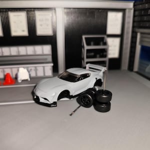 Könnte beinhalten: Ein graues Spielzeugauto mit schwarzen Rädern und einem silbernen Metallstab in einer Miniatur-Garagenszene. Dem Auto fehlen die Räder und es befindet sich in der Nähe eines Stapels schwarzer Reifen. Der Hintergrund umfasst ein Regal und Garagentore.