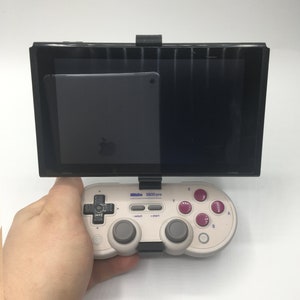May include: A white and black video game controller with purple buttons is held in a hand. A black tablet with a screen protector is attached to the controller with a black stand.
