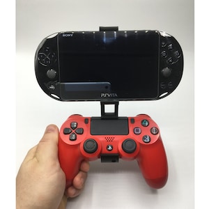 May include: A red PlayStation 4 controller with a black PlayStation Vita handheld console attached to it. The Vita is mounted on a black stand.