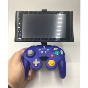 May include: A blue Nintendo Switch GameCube controller with a yellow, red, and green button layout. The controller is connected to a black Nintendo Switch console.