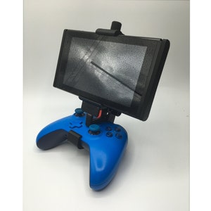 May include: A blue video game controller with a black phone or tablet holder attached to the top. The screen of the device is visible and shows a reflection of a window.