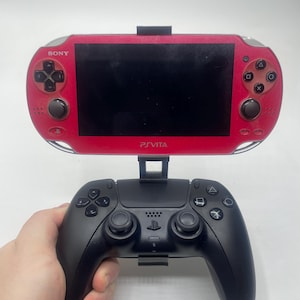May include: A red PS Vita handheld game console attached to a black controller. The console has the word "SONY" on the top left and "PSVITA" on the bottom. The controller has the PlayStation logo.