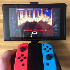 May include: A Nintendo Switch console with a black and red Joy-Con controller attached to the left side and a black and blue Joy-Con controller attached to the right side. The console is displaying the game "DOOM" with the text "Press to Start" on the screen.