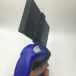 May include: A blue Nintendo GameCube controller attached to a black Nintendo Switch console with a black protective case. The Switch console is held vertically and the controller is held horizontally.
