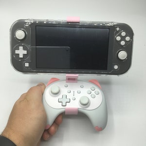 May include: A gray handheld gaming console with a clear protective case and a white controller. The controller has pink accents and a pink holder. The console and controller are connected with pink clips.