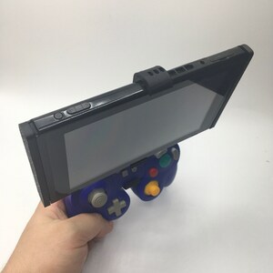 May include: A black Nintendo Switch console attached to a blue GameCube controller with a black 3D printed adapter. The Switch is in handheld mode and the screen is facing up.