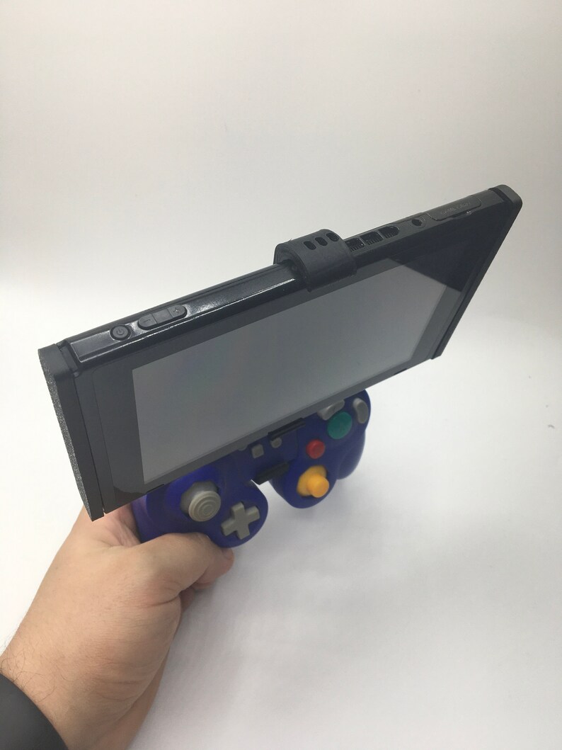 May include: A black Nintendo Switch console attached to a blue GameCube controller with a black 3D printed adapter. The Switch is in handheld mode and the screen is facing up.