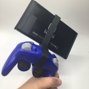 May include: A black Nintendo Switch console is attached to a blue and gray Nintendo GameCube controller with a 3D printed adapter. The Switch is held in a hand.