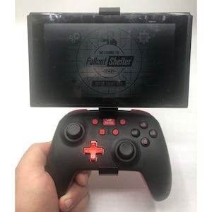 May include: A black and red video game controller with a screen displaying the Fallout Shelter game logo. The screen says "Welcome to Fallout Shelter" and "Enter Vault 420".