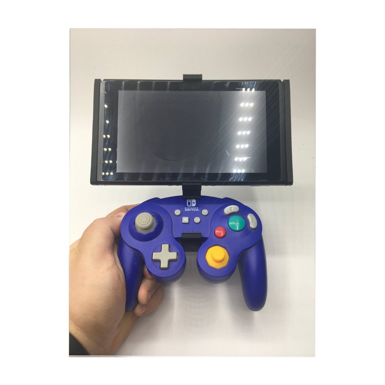 May include: A blue Nintendo Switch GameCube controller with a yellow, red, and green button layout. The controller is connected to a black Nintendo Switch console.