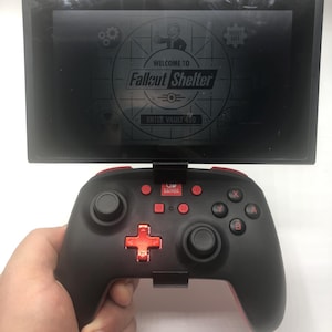 May include: A black Nintendo Switch console with a controller. The screen displays the words "Welcome to Fallout Shelter" and "Enter Vault 420". The controller is black with red accents and buttons.