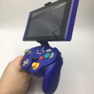 May include: A blue Nintendo Switch GameCube controller with a yellow button and gray buttons. The controller is connected to a purple Nintendo Switch with a black screen.