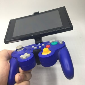 May include: A blue Nintendo Switch GameCube controller with a black Nintendo Switch console attached to it. The controller has a yellow button, a green button, and a gray button. The console has a black screen.