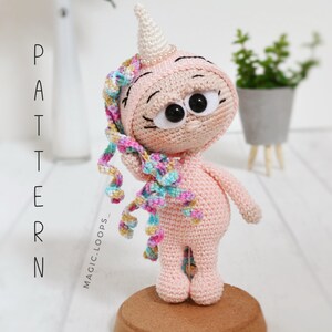 May include: A pink crocheted unicorn amigurumi toy with a rainbow mane and a white horn. The unicorn has large black eyes and a pink body. The toy is standing on a wooden base.