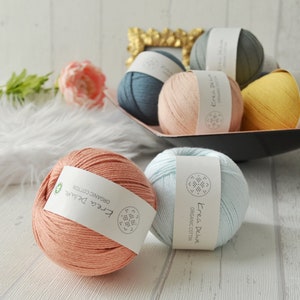 Könnte beinhalten: Fünf Knäuel Garn in verschiedenen Farben, darunter hellblau, rosa, gelb und orange. Das Garn ist mit "Free Deluxe Organic Cotton" gekennzeichnet.