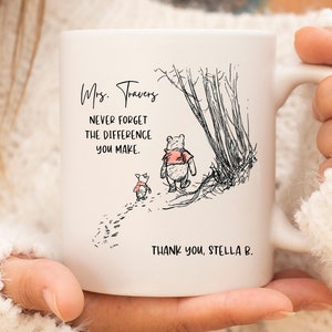 May include: A white ceramic mug featuring a black ink illustration of a bear and a small piglet walking near trees. The text reads: Mrs. Travers, NEVER FORGET THE DIFFERENCE YOU MAKE. THANK YOU, STELLA B. The design includes red accents on the clothing.