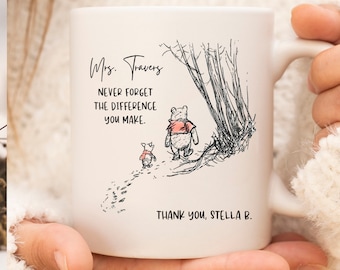 May include: A white ceramic mug featuring a black ink illustration of a bear and a small piglet walking near trees. The text reads: Mrs. Travers, NEVER FORGET THE DIFFERENCE YOU MAKE. THANK YOU, STELLA B. The design includes red accents on the clothing.
