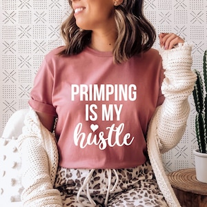 May include: A dusty rose t-shirt with the text "PRIMPING IS MY hustle" in white, paired with leopard print pants and a cream-colored cardigan. A cactus in a white pot sits on a wooden side table.