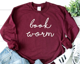 May include: A maroon long-sleeve sweatshirt with the words "book worm" in white script. The sweatshirt is paired with light blue jeans and white sneakers with black stripes. A small green plant is also in the image.
