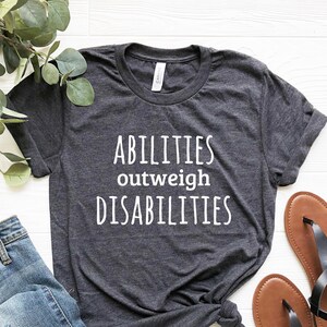 May include: A dark gray t-shirt with the text "ABILITIES outweigh DISABILITIES" in white lettering. The shirt is laid out on a white wooden surface with a pair of brown sandals and blue jeans.