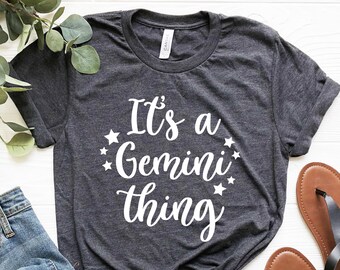 May include: Dark gray t-shirt with the text "It's a Gemini thing" in white script, surrounded by stars. The shirt is paired with blue jeans and brown sandals with black straps. A sprig of greenery is in the background.