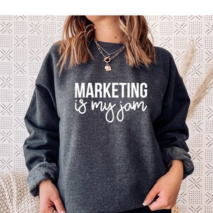 Puede incluir: Una sudadera gris oscuro con el texto "MARKETING is my jam" en letras blancas.
