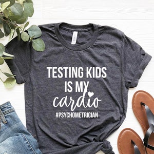 K&ouml;nnte beinhalten: Dunkelgraues T-Shirt mit wei&szlig;em Text: "TESTING KIDS IS MY cardio" mit einem Herzsymbol und dem Hashtag "#PSYCHOMETRICIAN". Das Shirt wird mit blauen Jeans und braunen Sandalen kombiniert.