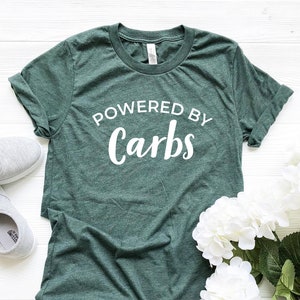 Może przedstawiać: Ciemnozielona koszulka z białym napisem "POWERED BY Carbs". Koszulka jest złożona, z szarymi trampkami i białymi kwiatami w tle. Koszulka jest wykonana z miękkiego materiału.