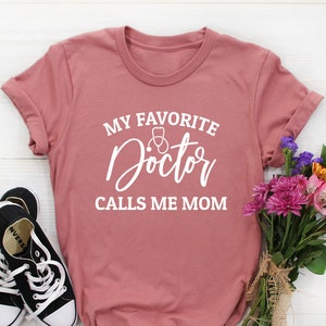 May include: A pink t-shirt with the text "My Favorite Doctor Calls Me Mom" in white lettering. The text includes a stethoscope and a heart graphic.