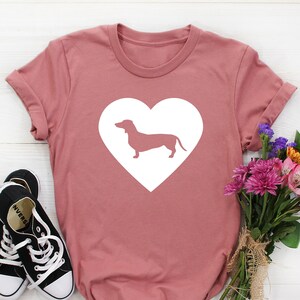 May include: Dusty rose t-shirt with a white heart and a dachshund silhouette. Black canvas trainers with white laces are on the left. A bouquet of colourful flowers tied with twine is on the right. The shirt has rolled-up sleeves.