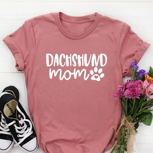 以下が含まれることがあります： ダックスフントママと白文字で書かれた、くすんだピンクのTシャツ。ママの隣には白い肉球のプリントがあります。