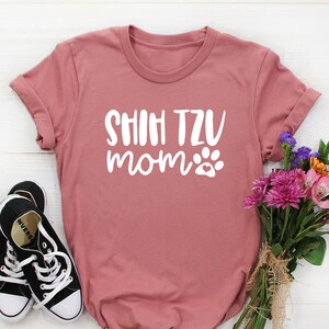 May include: A dusty rose t-shirt with the text "SHIH TZU MOM" in white with a paw print. The shirt is laying on a white wooden surface with a bouquet of flowers and a pair of black sneakers.