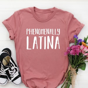 May include: A dusty rose t-shirt with the words "PHENOMENALLY LATINA" in white block letters. Black and white canvas sneakers are to the left of the shirt. A bouquet of colorful flowers is to the right.